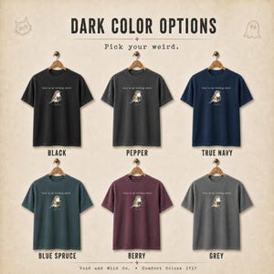 May include: Six short-sleeve t-shirts in black, pepper, true navy, blue spruce, berry, and grey. Each shirt features a small graphic of a bird holding a letter M and the text this is my birding shirt. The image includes the text DARK COLOR OPTIONS.