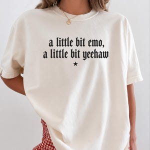 May include: An off-white short-sleeve t-shirt featuring black text printed on the chest that reads, a little bit emo, a little bit yeehaw, with a small black star symbol centered directly below the text. The shirt has a crew neckline.