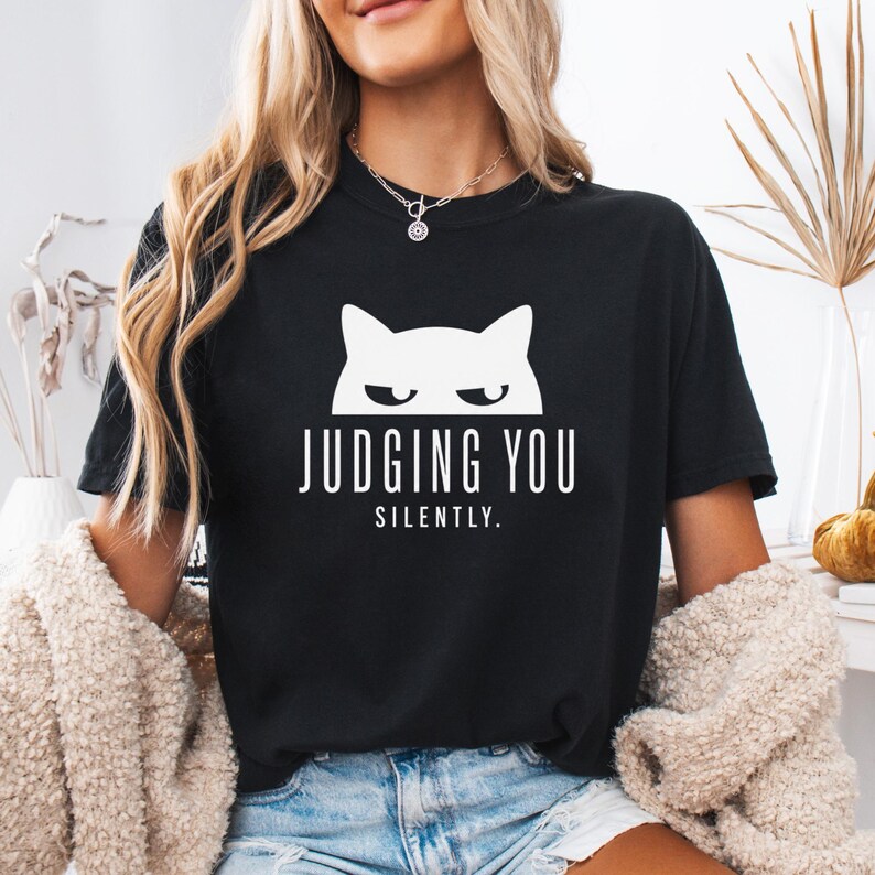 May include: Black t-shirt with a white graphic of a cat's face and the words "JUDGING YOU SILENTLY." The shirt is a crew neck style.