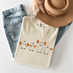 Pode incluir: Uma t-shirt cor de creme com um gráfico de quatro fantasmas brancos a levantar halteres e uma barra de pesos cor de laranja. A t-shirt está sobre calças de ganga azul-claras sob um chapéu bege de aba larga. Uma corrente dourada fina é visível.