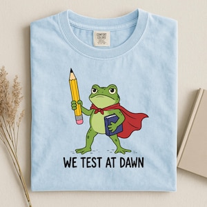 May include: A light blue t-shirt featuring a graphic of a green frog wearing a red cape, holding a yellow pencil and a blue book. Below the frog, the text reads WE TEST AT DAWN. The shirt has a Comfort Colors brand label at the inner neckline.