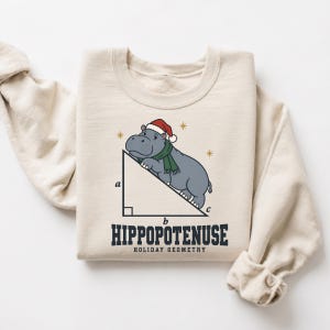 May include: A beige crewneck sweatshirt featuring a graphic of a grey hippopotamus wearing a red Santa hat and green scarf, positioned on the hypotenuse of a right triangle labelled with a, b, and c. Text reads HIPPOPOTENUSE HOLIDAY GEOMETRY.
