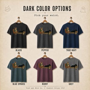 May include: Six short-sleeve t-shirts in black, pepper, true navy, blue spruce, berry, and grey. Each shirt features a graphic of a cat sitting in a chair reading and a black cat lying on a couch. Text reads: DARK COLOR OPTIONS, Pick your weird.
