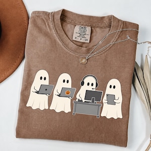 Könnte beinhalten: Ein braunes Kurzarm-T-Shirt mit einer Grafik von vier weißen Geistern, die Technologie nutzen. Ein Geist benutzt einen Laptop, ein anderer einen Laptop mit Kürbisaufkleber, einer sitzt an einem Schreibtisch mit Computer und Headset, und einer hält ein Tablet.