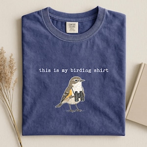 May include: A blue crew neck t-shirt featuring a graphic of a bird holding black binoculars. The text "this is my birding shirt" is printed in white above the bird. The shirt has a Comfort Colors brand label on the inside of the collar.
