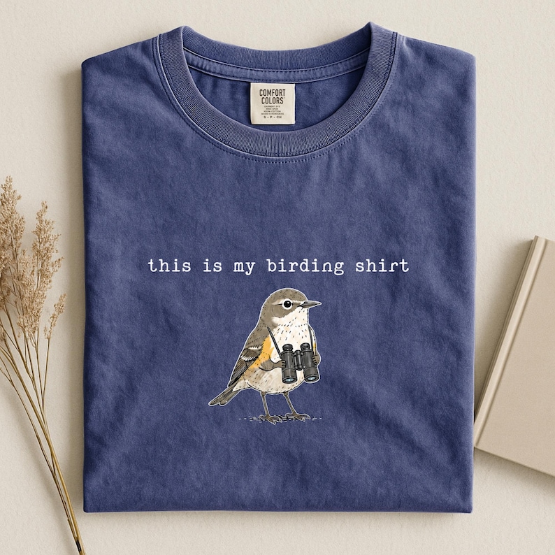 May include: A blue crew neck t-shirt featuring a graphic of a bird holding black binoculars. The text "this is my birding shirt" is printed in white above the bird. The shirt has a Comfort Colors brand label on the inside of the collar.