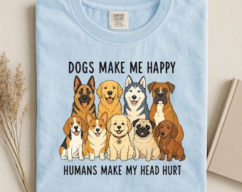May include: A light blue t-shirt featuring a graphic illustration of nine different dog breeds arranged in two rows. Above the dogs, the text reads "DOGS MAKE ME HAPPY" and below the dogs, the text reads "HUMANS MAKE MY HEAD HURT" in black lettering.