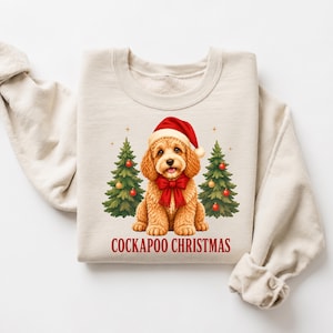 Puede incluir: Una sudadera beige de cuello redondo con el gráfico de un perro Cockapoo de color marrón dorado que lleva un gorro de Papá Noel rojo y un lazo rojo en el cuello. El perro está centrado entre dos árboles de Navidad verdes con adornos rojos. Texto: COCKAPOO CHRISTMAS.