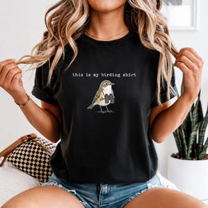 May include: A black short-sleeved t-shirt featuring a graphic of a small bird wearing binoculars around its neck. Above the bird, the text reads, "this is my birding shirt" in a white, typewriter-style font. The shirt has a crew neckline.