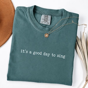 Puede incluir: Una camiseta de manga corta color verde azulado con cuello redondo y el texto blanco "it's a good day to sing" impreso en el pecho. La camiseta cuenta con una etiqueta de la marca Comfort Colors en el cuello y un collar con dije dorado.