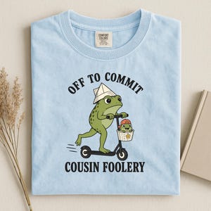 May include: A light blue t-shirt featuring a graphic of a green frog wearing a paper hat, riding a black scooter with a small frog in a basket. The text reads OFF TO COMMIT COUSIN FOOLERY. The shirt has a Comfort Colors brand label at the neck.