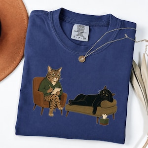 May include: A navy blue t-shirt featuring a graphic of a tabby cat in a green sweater sitting in a brown armchair, holding a notepad and pencil, while a black cat lies on a brown chaise lounge nearby with a tissue box on the floor.