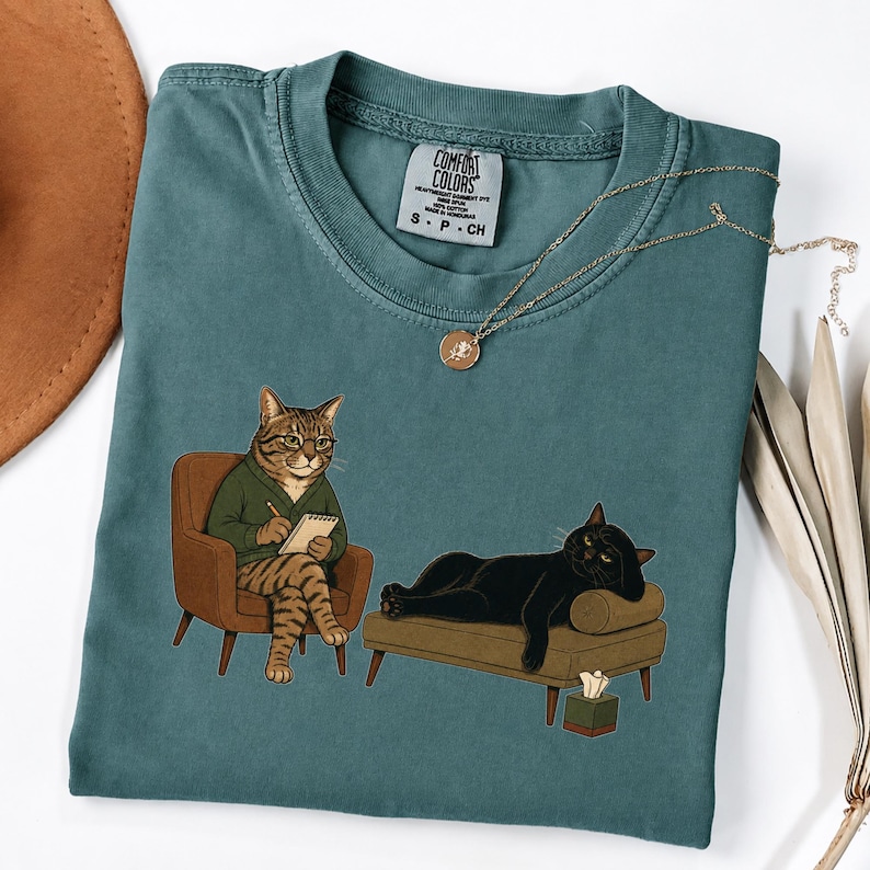 May include: A teal crewneck t-shirt featuring a graphic of a tabby cat therapist in a green sweater sitting in a chair with a notepad, and a black cat lying on a couch. The shirt has a Comfort Colors label and a gold-toned pendant necklace.