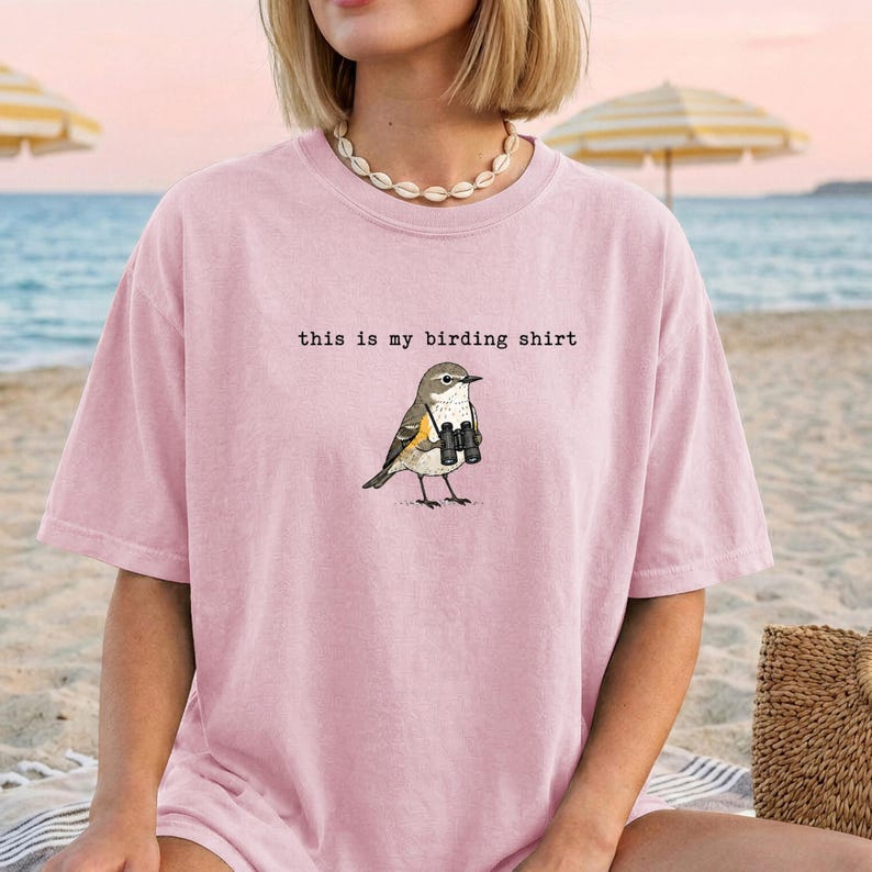 May include: A light pink short-sleeved t-shirt featuring a graphic of a small bird wearing binoculars around its neck. Above the bird, the text reads "this is my birding shirt" in a simple, black, lowercase typewriter-style font.