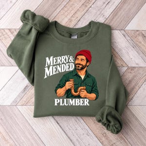 May include: An olive green crewneck sweatshirt featuring a graphic of a person with a beard wearing a red beanie and a green shirt, holding a copper pipe fitting. The text reads MERRY & MENDED PLUMBER in white, bold, sans-serif lettering.