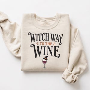 Könnte beinhalten: Ein beigefarbenes Sweatshirt mit Rundhalsausschnitt und dem schwarzen und orangefarbenen Schriftzug "WITCH WAY TO THE WINE" auf der Vorderseite. Unter dem Text befindet sich eine Grafik eines Weinglases mit Rotwein und einem kleinen Hexenhut.