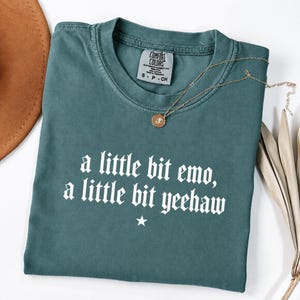 May include: A teal short-sleeved t-shirt featuring the white text 'a little bit emo, a little bit yeehaw' with a small star symbol below the lettering. The shirt has a crew neckline and a Comfort Colors brand label inside the collar.