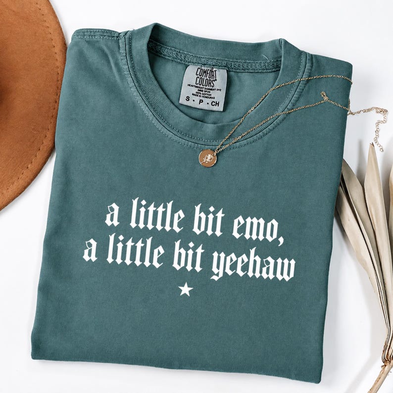 May include: A teal short-sleeved t-shirt featuring the white text 'a little bit emo, a little bit yeehaw' with a small star symbol below the lettering. The shirt has a crew neckline and a Comfort Colors brand label inside the collar.