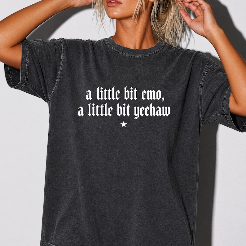 May include: A charcoal grey short-sleeved t-shirt with a washed-out, vintage-style finish. The front features white text in a gothic-inspired font that reads, a little bit emo, a little bit yeehaw, centered above a small, simple white star icon.