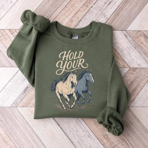 May include: An olive green crewneck sweatshirt featuring a graphic print of two running horses, one cream and one dark blue, with the text "HOLD YOUR" in a cream-colored script font positioned above the horses on the front of the garment.