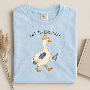 Op de afbeelding: Een lichtblauw t-shirt met ronde hals en een afbeelding van een witte gans met een gele veiligheidshelm. De gans draagt een blauwe opgerolde bouwtekening en een geodriehoek. Boven de gans staat de tekst OFF TO ENGINEER in zwarte letters.