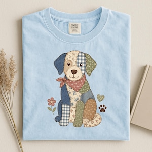 Può includere: Una t-shirt azzurro chiaro con la grafica di un cane disegnato con vari motivi patchwork nei toni del blu, verde, marrone e crema. Il cane indossa una bandana rossa ed è circondato da un piccolo fiore, un cuore e un'impronta di zampa.