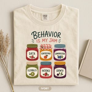 Puede incluir: Una camiseta de color marfil con el texto BEHAVIOR IS MY JAM y seis frascos ilustrados etiquetados como DATA, REINFORCEMENT, COMMUNICATION, FLEXIBILITY, PATIENCE y GROWTH, cada uno decorado con ilustraciones de frutas como frambuesas, manzanas y arándanos.