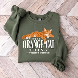 May include: An olive green crewneck sweatshirt featuring a graphic of a reclining orange tabby cat. Below the cat, white text reads: IT'S AN ORANGE CAT THING YOU WOULDN'T UNDERSTAND. The sweatshirt has long sleeves with ribbed cuffs and a ribbed neckline.