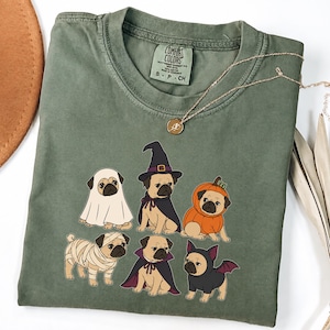 Könnte beinhalten: Ein olivgrünes T-Shirt mit einem Grafikdruck von sechs Möpsen in Halloween-Kostümen, darunter ein Geist, eine Hexe, ein Kürbis, eine Mumie, ein Vampir und eine Fledermaus. Das T-Shirt hat einen Rundhalsausschnitt und ein Comfort Colors Markenetikett.
