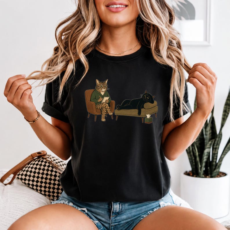 May include: A black short-sleeve t-shirt featuring a graphic illustration of a tabby cat in a green sweater sitting in a chair writing on a notepad, while a black cat lies on a couch nearby with a tissue box placed on the floor beside it.