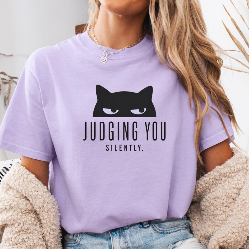 May include: A light purple t-shirt with a black cat graphic above the words "JUDGING YOU SILENTLY." The cat graphic features a simple silhouette with visible eyes. The shirt has short sleeves and a crew neck.