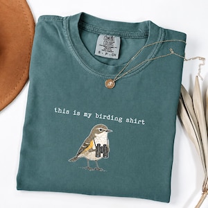 May include: A teal crewneck t-shirt with a graphic of a small bird holding black binoculars. The text "this is my birding shirt" is printed in white above the bird. The shirt features a Comfort Colors brand label inside the neckline.