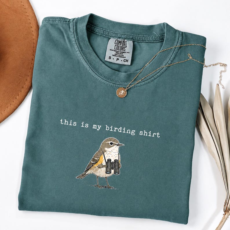 May include: A teal crewneck t-shirt with a graphic of a small bird holding black binoculars. The text "this is my birding shirt" is printed in white above the bird. The shirt features a Comfort Colors brand label inside the neckline.