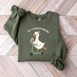Puede incluir: Una sudadera de cuello redondo color verde oliva con el gráfico de un pato blanco con pico y patas naranjas, con un lazo rosa, montado en un patinete azul verdoso mientras sostiene un café helado. El texto superior dice OFF TO REGULATE EMOTIONS.