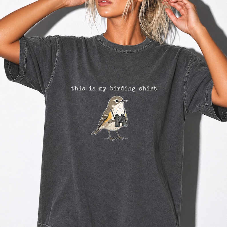 May include: A charcoal gray short-sleeve t-shirt featuring a graphic of a small brown bird holding black binoculars. Above the bird, the text reads, "this is my birding shirt" in a simple white typewriter-style font. The fabric has a washed-out look.