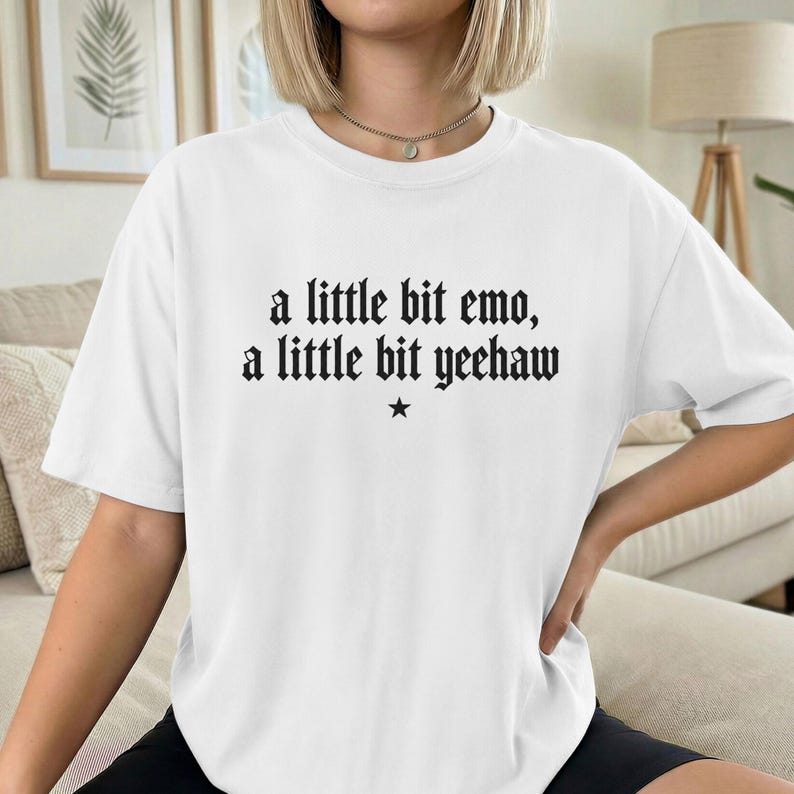 May include: A white short-sleeved t-shirt featuring black text printed on the chest that reads, a little bit emo, a little bit yeehaw, with a small black star symbol centered directly below the text in a gothic-style font.