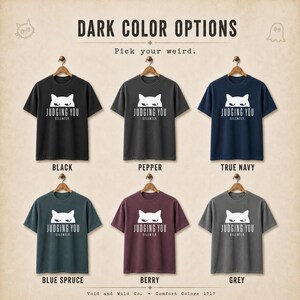 May include: A display of six t-shirts in various dark colors, each with a cat graphic and the text "JUDGING YOU SILENTLY." Colors include black, pepper, true navy, blue spruce, berry, and grey. The shirts are hanging on wooden hangers.