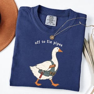 Könnte beinhalten: Ein marineblaues T-Shirt mit einer Grafik einer weißen Gans, die eine Werkzeugweste trägt und einen Schraubenschlüssel hält. Der Text über der Gans lautet "off to fix pipes". Das T-Shirt hat ein Comfort Colors Markenetikett im Kragen.