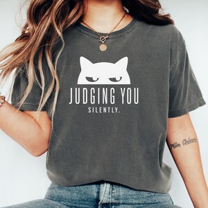 May include: Dark gray t-shirt with a white cat graphic and the text "JUDGING YOU SILENTLY." The shirt has a crew neck and short sleeves. The cat graphic features a minimalist design with a frowning expression.