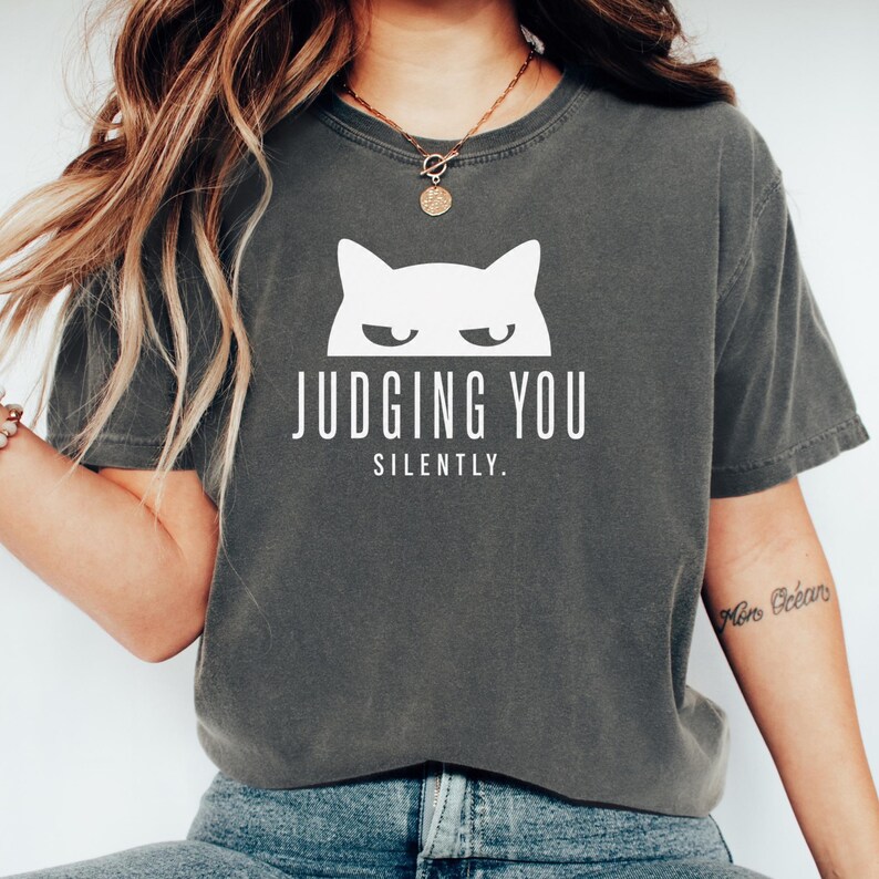 May include: Dark gray t-shirt with a white cat graphic and the text "JUDGING YOU SILENTLY." The shirt has a crew neck and short sleeves. The cat graphic features a minimalist design with a frowning expression.