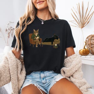 May include: A black t-shirt featuring a graphic illustration of a tabby cat in a green sweater sitting in a chair with a notepad, observing a black cat lying on a chaise lounge. A small tissue box is placed on the floor near the lounge.