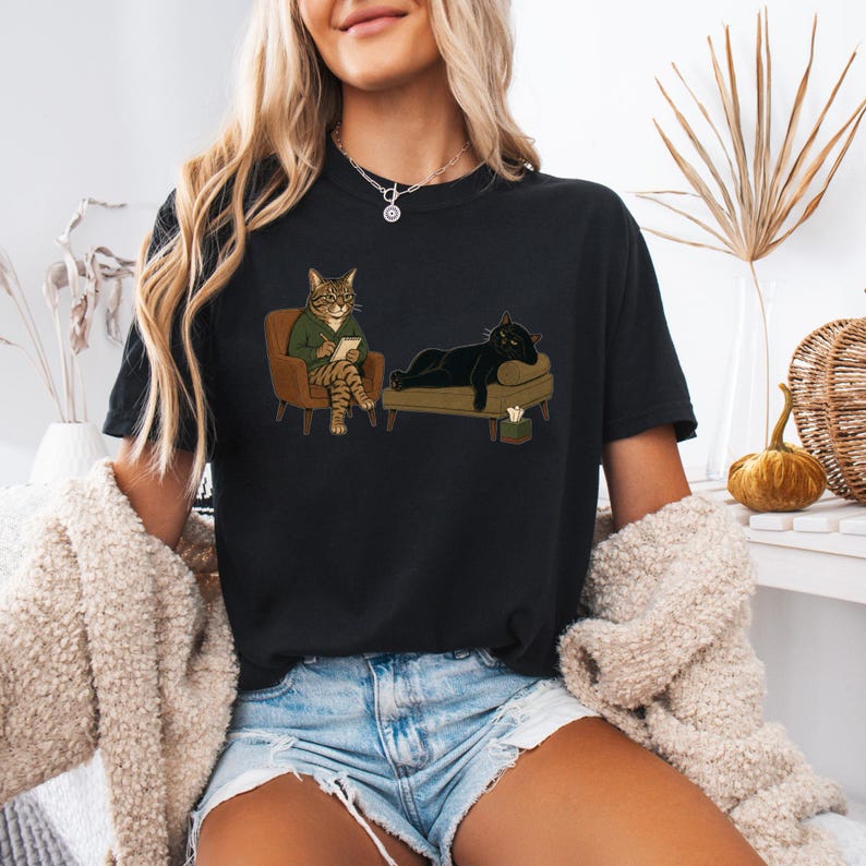 May include: A black t-shirt featuring a graphic illustration of a tabby cat in a green sweater sitting in a chair with a notepad, observing a black cat lying on a chaise lounge. A small tissue box is placed on the floor near the lounge.