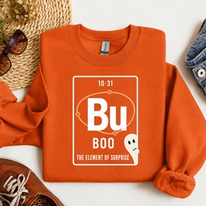 Peut inclure: Un sweat-shirt orange à manches longues avec un motif blanc rappelant un élément du tableau périodique. Le graphique affiche le texte 10-31, les lettres Bu, le mot BOO et le texte THE ELEMENT OF SURPRISE, avec une petite illustration de fantôme.