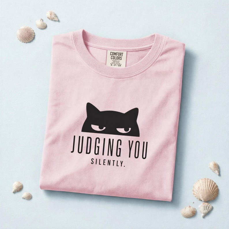 May include: A light pink t-shirt with a black cat graphic and the text "JUDGING YOU SILENTLY." The shirt is folded neatly and surrounded by seashells. The shirt has a crew neck and short sleeves.