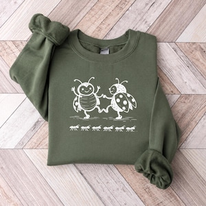Op de afbeelding: Een olijfgroene sweater met ronde hals en een witte grafische print van twee gestileerde lieveheersbeestjes die samen dansen, geplaatst boven een horizontale rij van zes kleine mieren die in een rij over de borst lopen.
