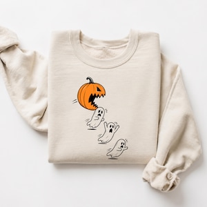 Puede incluir: Una sudadera de cuello redondo en color beige con un estampado gráfico de una calabaza naranja con la boca abierta persiguiendo a tres pequeños fantasmas blancos asustados en la parte frontal de la prenda de manga larga.