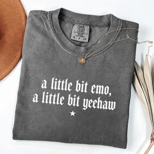 May include: A charcoal grey short-sleeve t-shirt with a crew neckline. The shirt features white text that reads, a little bit emo, a little bit yeehaw, with a small white star centered below the text. A gold-toned necklace rests on the shirt.