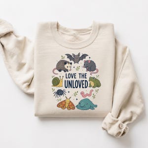 Peut inclure: Un sweat-shirt à col rond beige avec une impression graphique colorée représentant divers animaux, dont une chauve-souris, un opossum, un rat, un escargot, une araignée, un papillon, un ver, une grenouille et un blobfish, avec le texte "LOVE THE UNLOVED".
