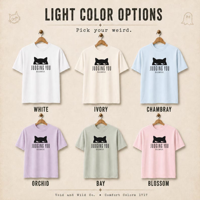 May include: A display of six short-sleeved t-shirts in various light colors, including white, ivory, and pink. Each shirt features a black cat graphic with the text "JUDGING YOU SILENTLY." The shirts are hanging on wooden hangers.