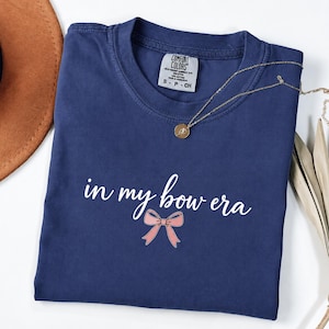 May include: A navy blue short-sleeved t-shirt featuring the white text "in my bow era" printed above a small pink bow graphic. The shirt has a crew neckline and a Comfort Colors brand label inside the collar, indicating a size small.